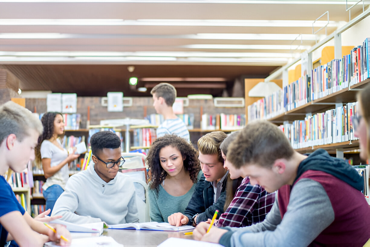 Diverse students studying together at a library table with bookshelves in the background.